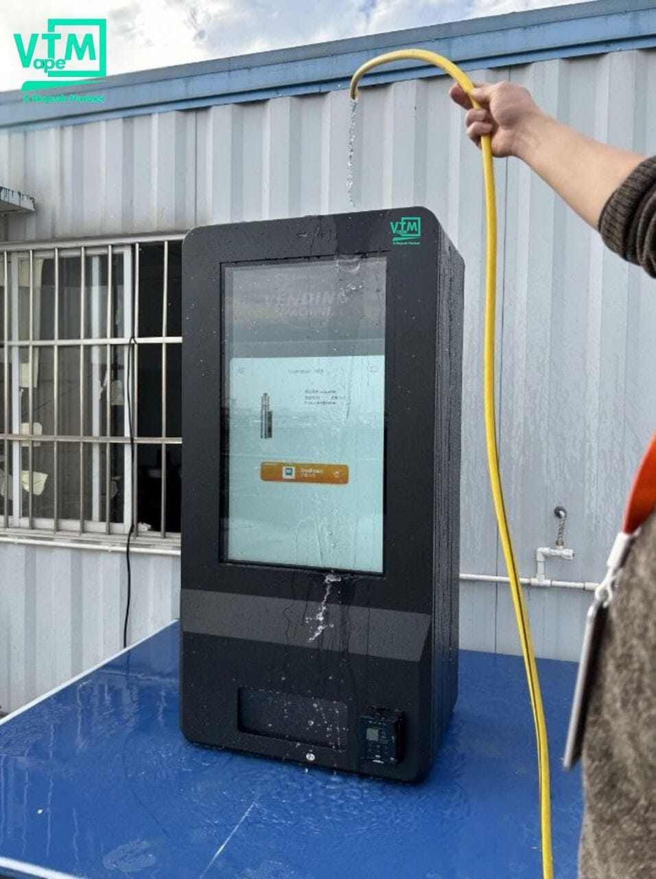 Outdoor VTM Vape vending machine undergoing waterproof test with water being sprayed from a hose, demonstrating weatherproof capabilities